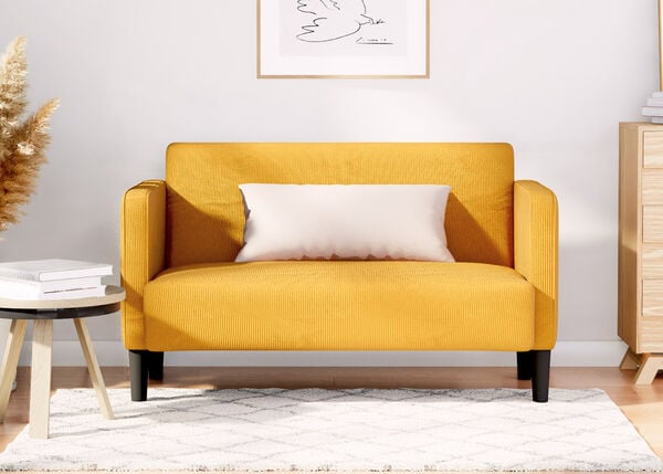 Yellow two-seater sofa with white pillow, next to wooden side table and wooden dresser, on patterned rug in a styled living room.