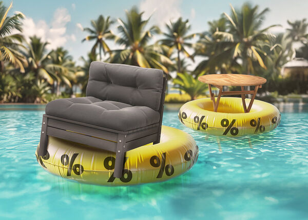 A dark gray cushioned chair and a round wooden outdoor table each floating on yellow inflatable rings with black percent signs, in a sunny pool surrounded by palm trees.