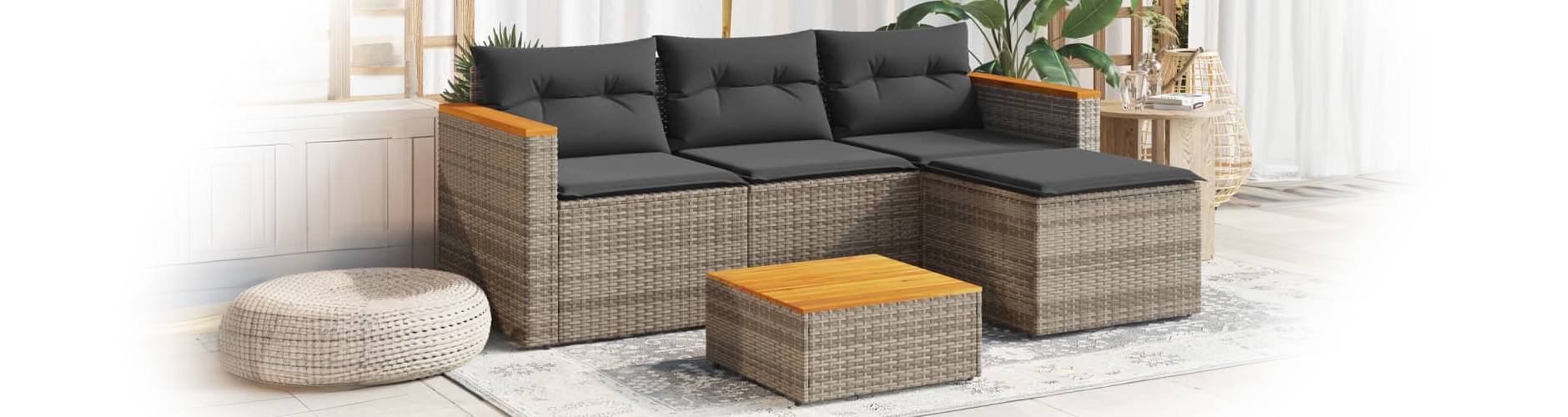Modular rattan outdoor sofa with dark gray cushions, matching coffee table on rug in a styled terrace. Set against white curtains and trellis with plants.