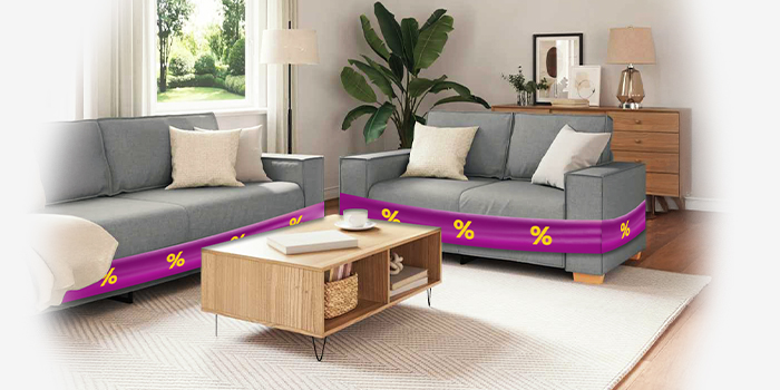 Two grey sofas wrapped in purple promotional bands showing yellow discount icons and wooden coffee table, in a styled living room. Click to shop the collection.
