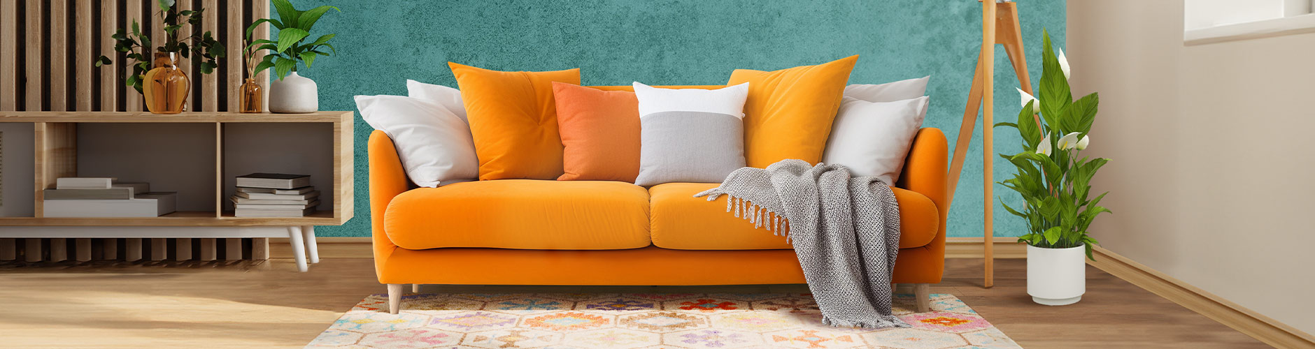 A modern living room with an orange plush sofa decorated with orange and white cushions and a gray throw blanket, a green plant beside it, and an open-shelf TV stand holding books and topped with another green plant..