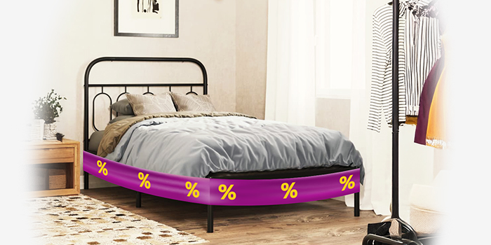 Black metal bed frame with grey duvet and pillows, wrapped in a purple promotional band showing yellow discount icons, next to wooden bedside table, in a styled bedroom. Click to shop the collection.